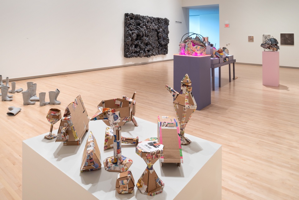 Overview of gallery displaying cardboard objects on a plinth, grey rubber boots, tables of objects lit with neon lights underneath, and a large dark wall-hung sculpture.