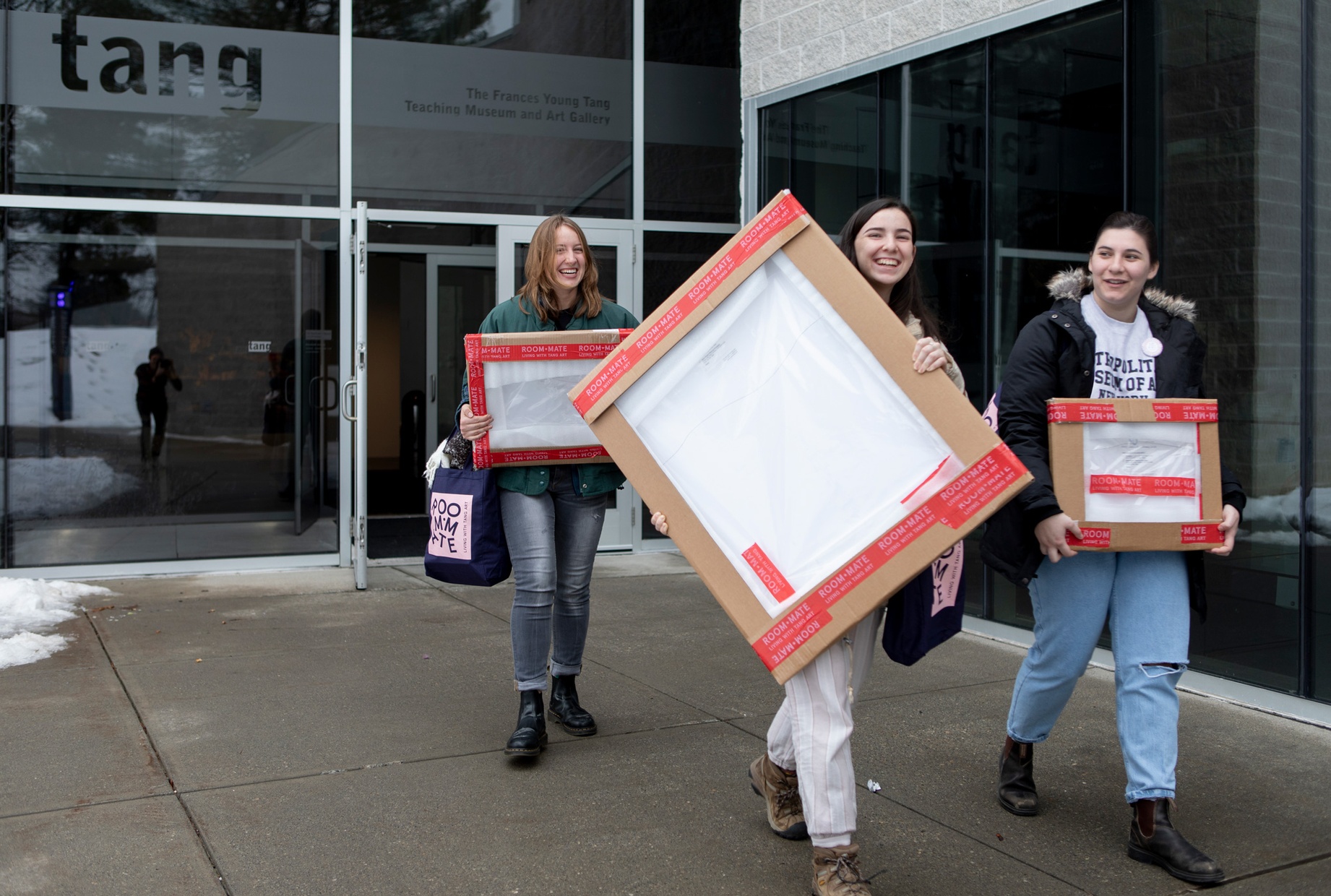Three students walking together each carrying a packaged artwork leave the Tang.