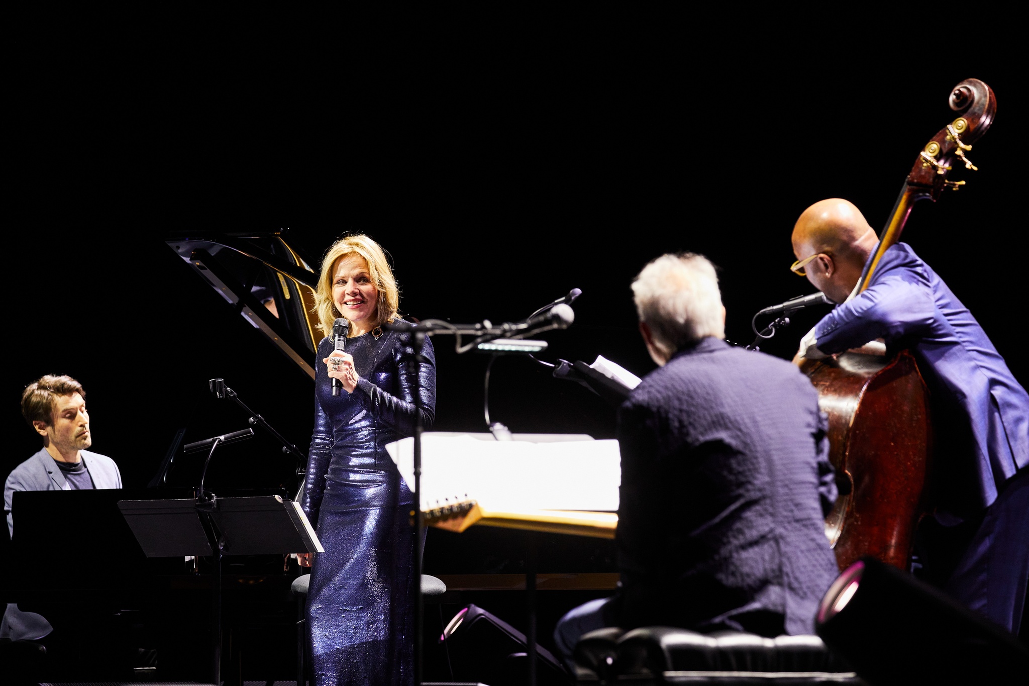 Four musicians on stage, a man seated at a piano, a woman in a purple dress holding a microphone, and two men seated with a bass and a guitar respectively
