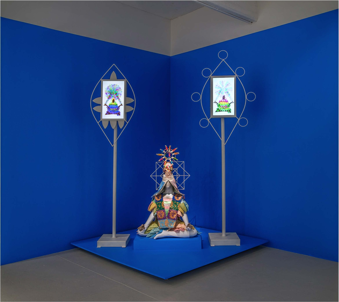 A humanoid figure in a colorful, petal-like robe and star-like headdress sits crosslegged on a blue platform flanked by two gray stands holding drawings of similar figures as the seated one.