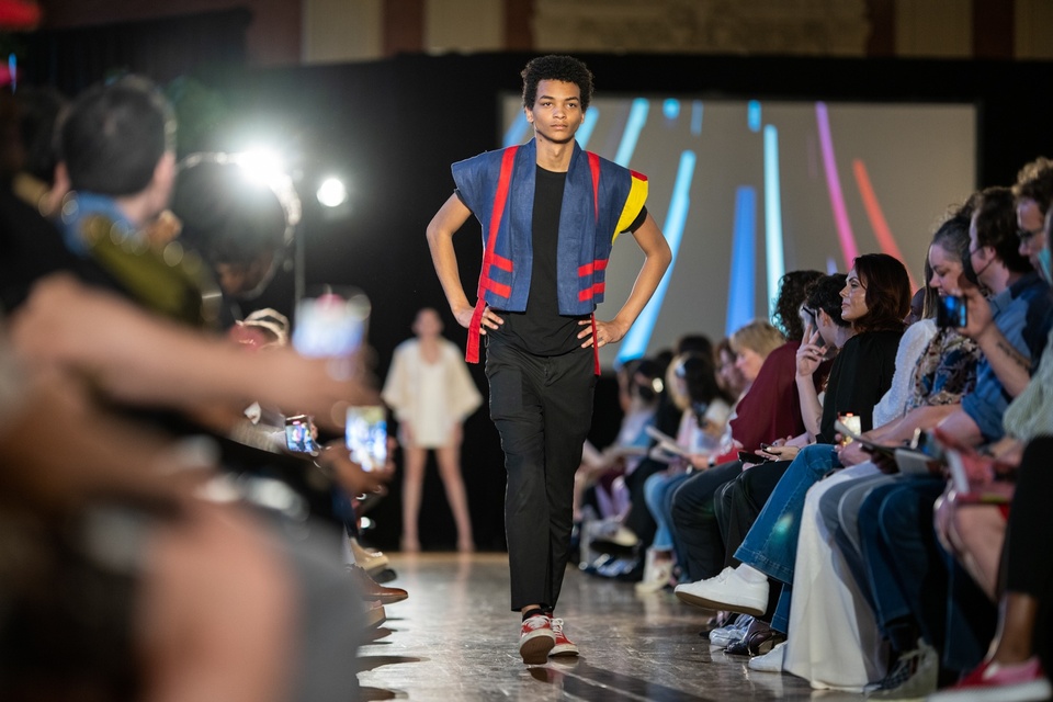 A model walking down the runway in a black pants and shirt with a blue denim stiff trapeze shaped jacket