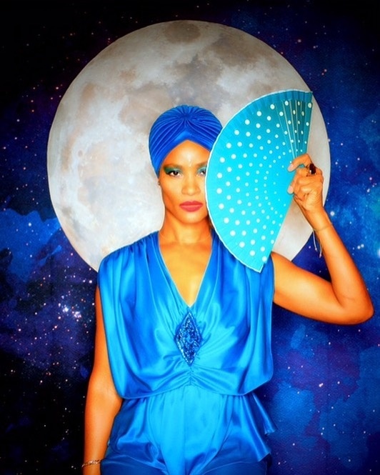 A portrait of DJ Jennifly in a vibrant blue jumpsuit and headscarf posing in front of a background showing the moon. She holds a light blue fan spread in front of the side of her face. 