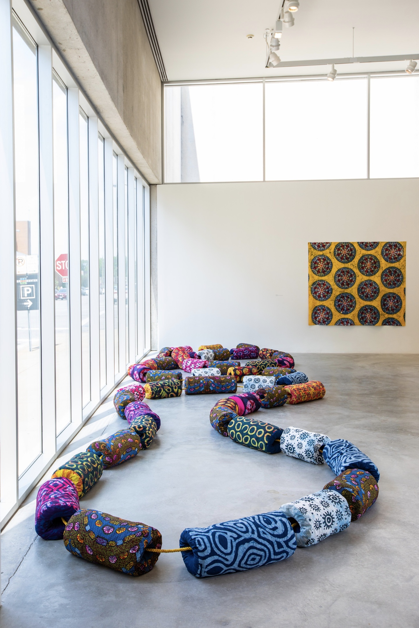 Photo of a sculptural installation in a white box gallery, with floor-to-ceiling windows to the left and a bank of high windows on the back wall. In the foreground, on the floor, is a sculptural installation of large, fabric-covered cylinders, strung together like a necklace, roughly shaped like an 8. On the back wall is an artwork with a golden background and circles. 