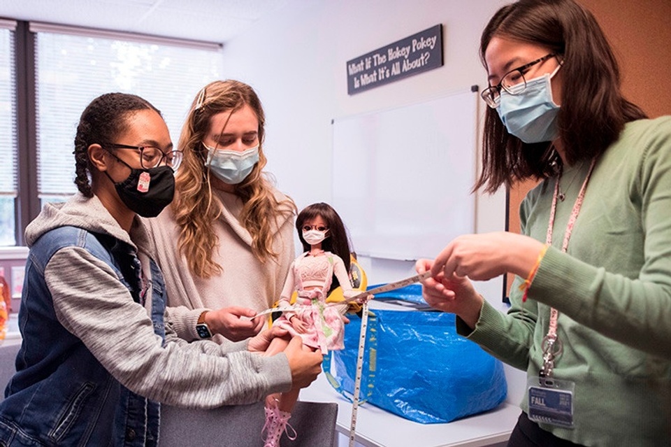 Three people wearing Covid masks gather around a small doll. One person is holding and looking at a measuring tape.