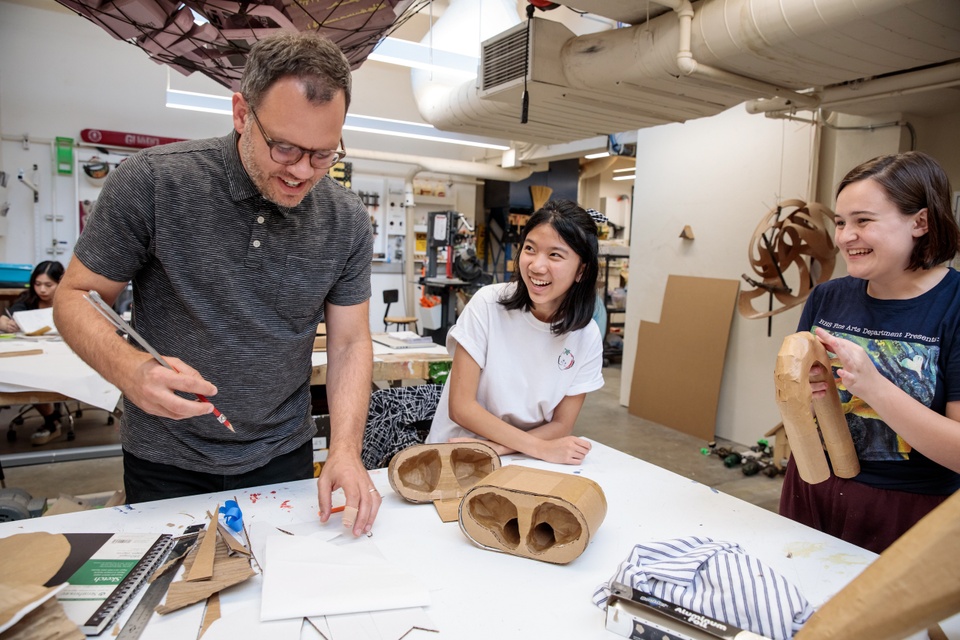 Three people stand around a worktable looking at a giant cardboard model of a AirPods case and associated plan drawings, laughing.