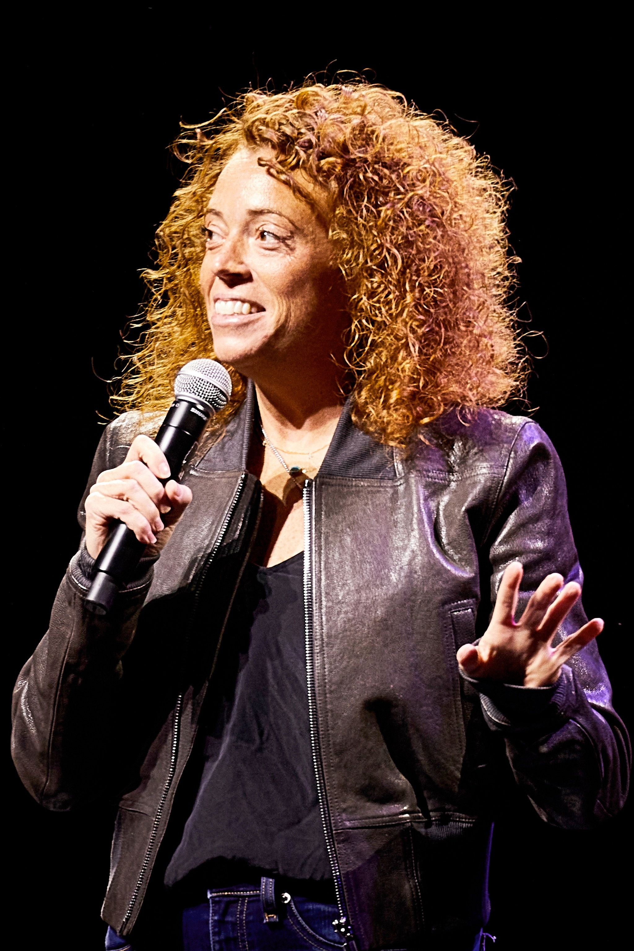 A close-up photo of comedian Michelle Wolf performing on stage. She has red curly hair, wears a leather bomber jacket, and holds a mic below her mouth. 