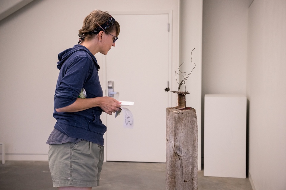 A visitor examines a small wire sculpture attached to an anvil on top of a concrete pedestal