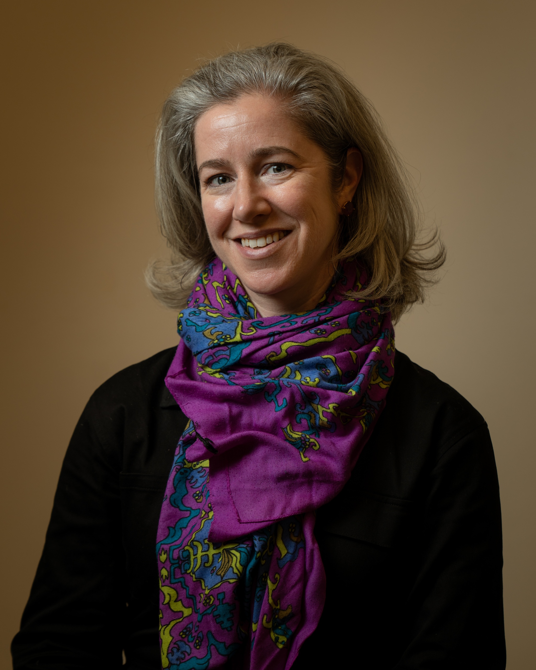 A portrait of Andrea Salvatore against a neutral brown background. Andrea has neck-length silvery gray hair and smiles broadly at us. She wears a voluminous scarf that is deep purple and blue. 