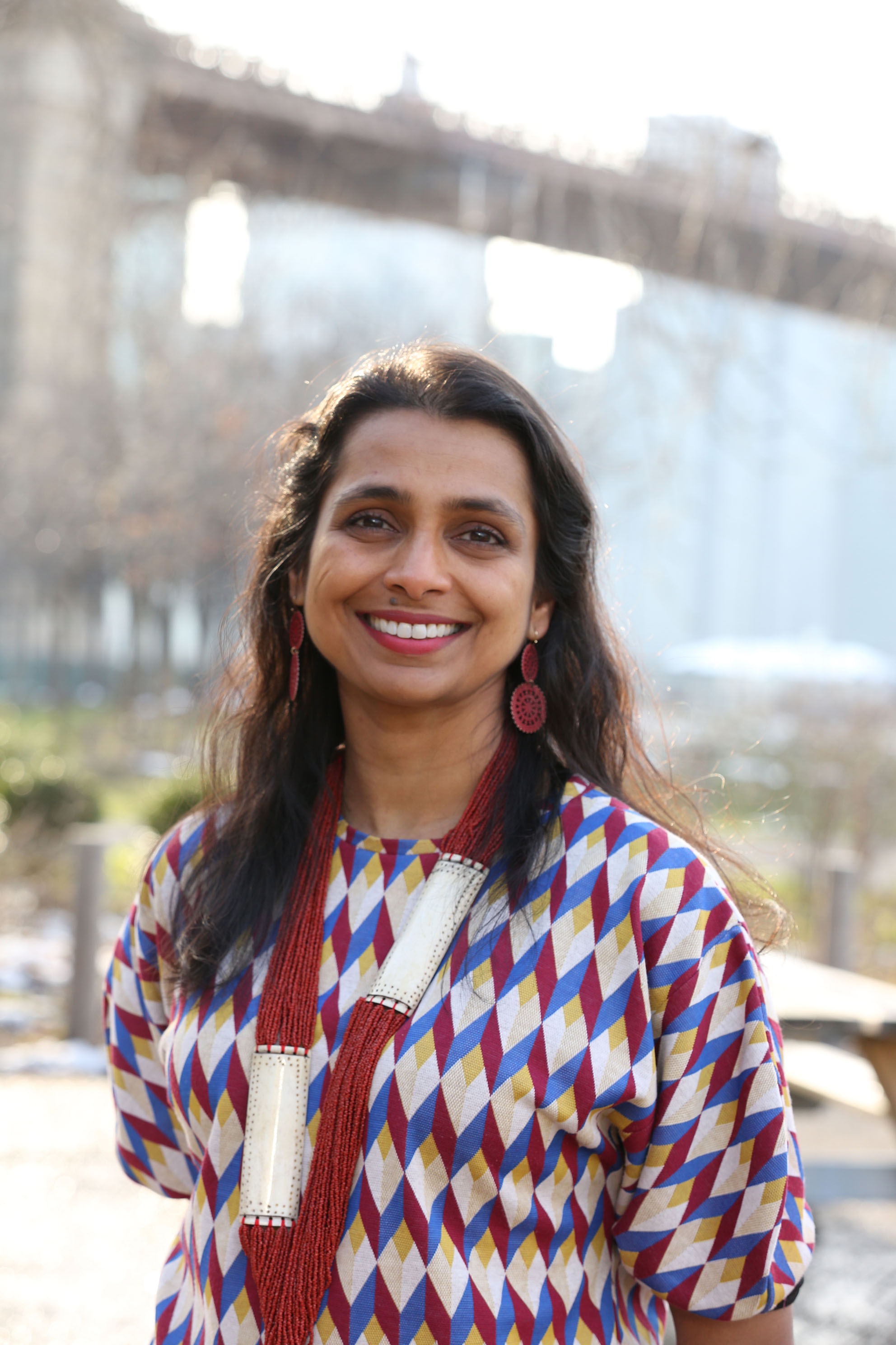A photo of Prerana Reddy standing against the blurry background of trees and a tall bridge. Reddy wears a geometric-print shirt, red earrings, and a long, red beaded necklace.