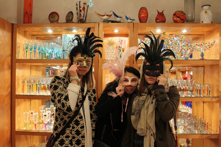 Three people in a glass shop holding ornate carnival masks over their eyes