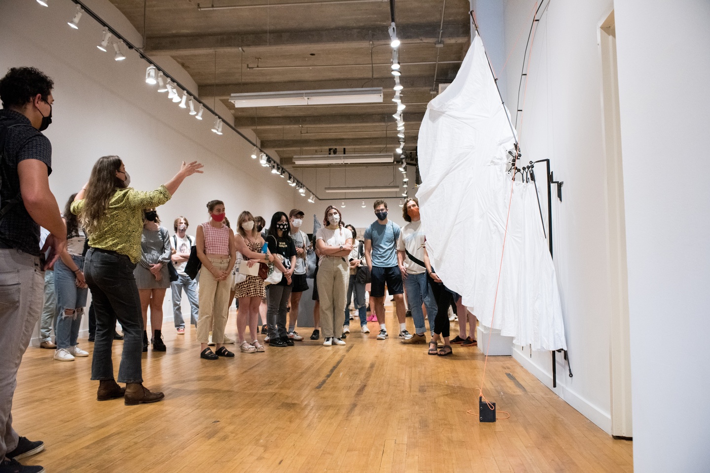 Artist delivers a gallery talk to visitors at a small gallery space. A white wing-like sculpture is seen in the foreground.