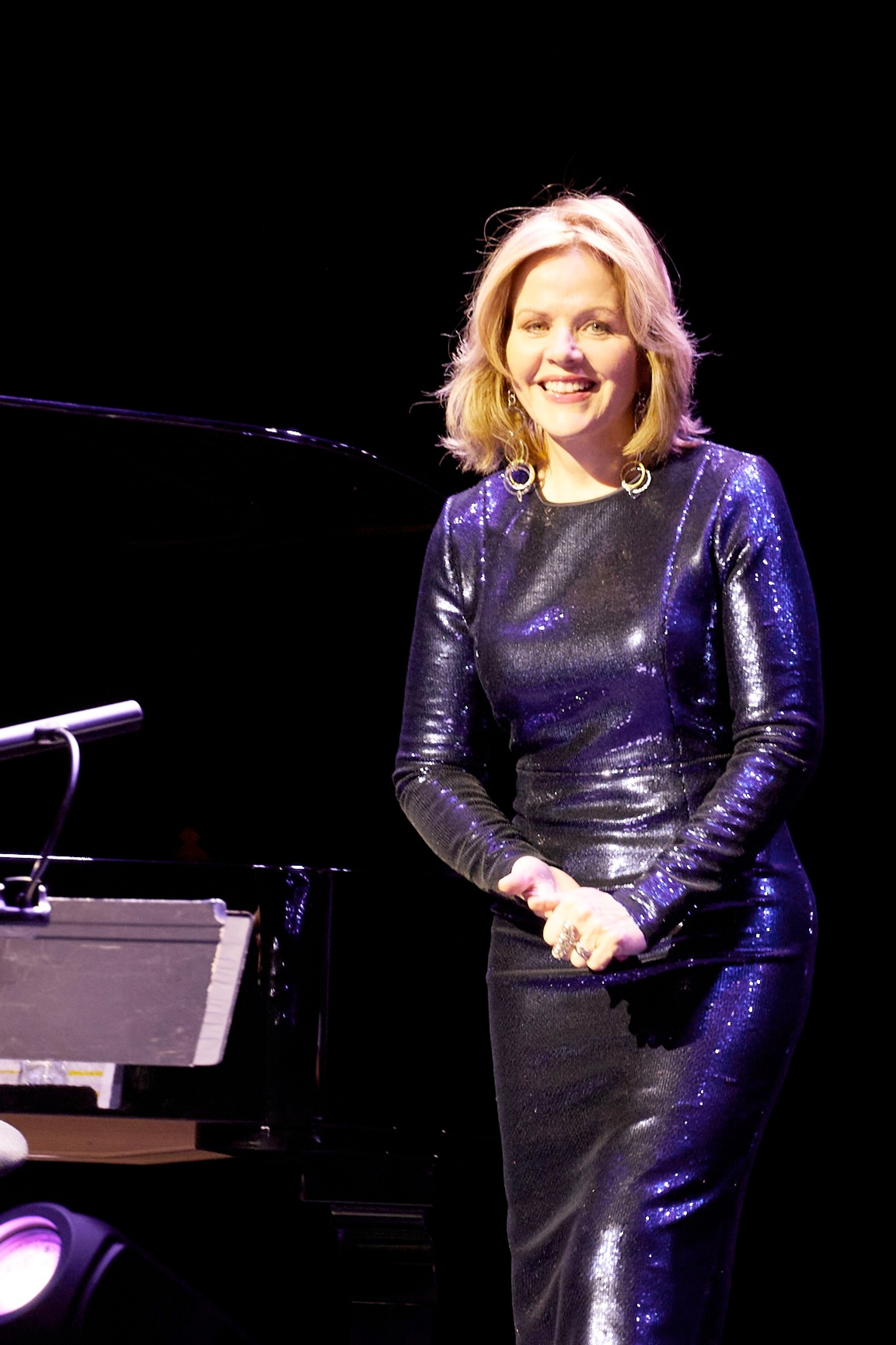 Soprano Renée Fleming smiling on stage with her hands clasped in front of her. She has blond hair that falls to her shoulders and wears a dress covered in purple sequins that shine in the stage light. 