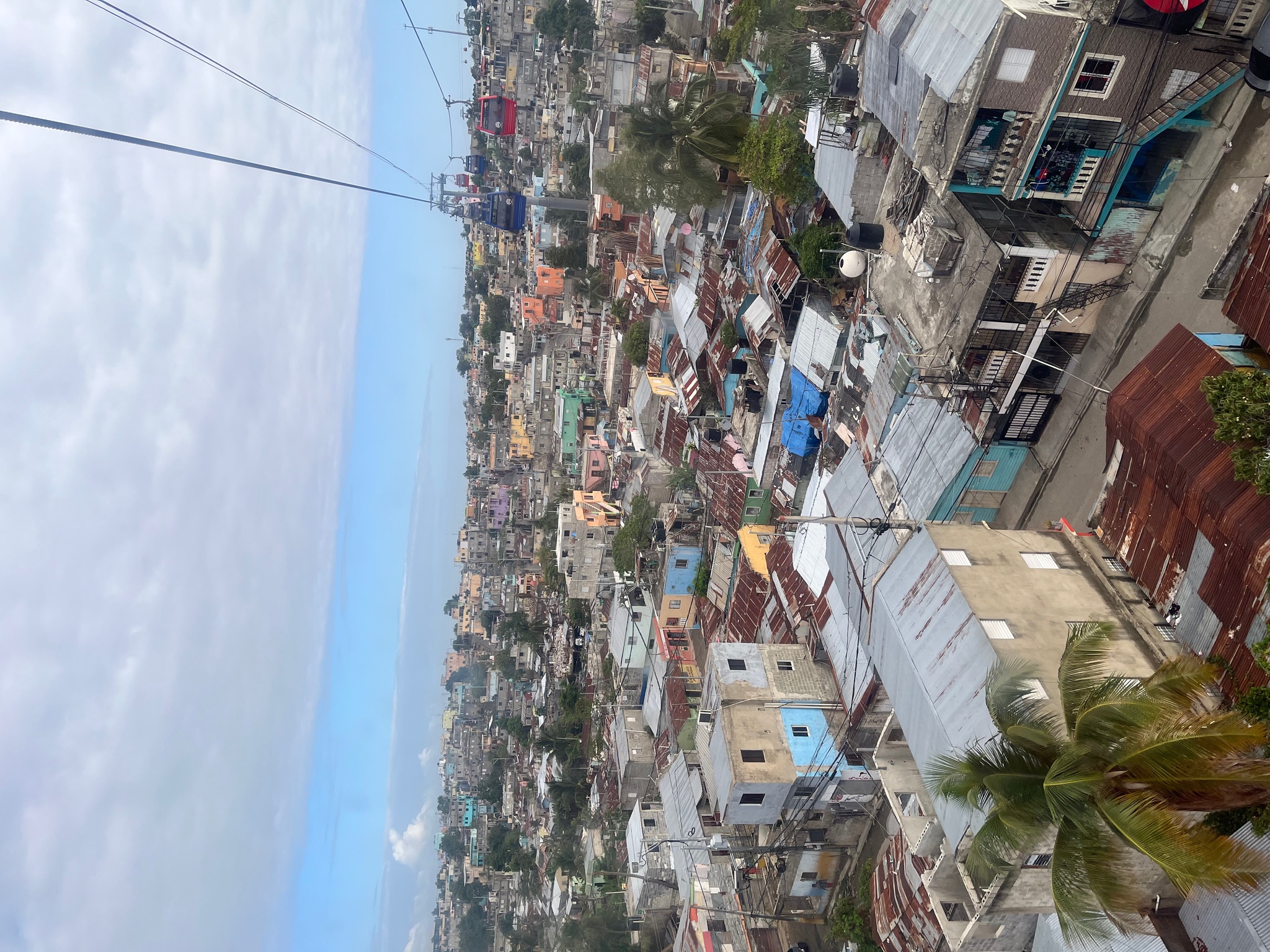 A neighborhood in the Dominican Republic seen from above, taken from the gondola of an aerial tram. The buildings are painted soft blue and pinks. On the horizon is a strip of light blue sky with light gray clouds above it. 