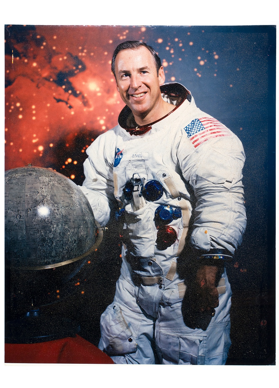 Photographic portrait of a man in a space suit smiling with one hand on a globe and an image of a galaxy in the background.