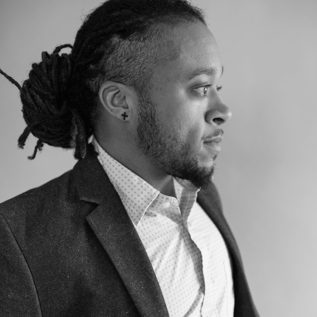 A Black man seen in profile. He wears a black blazer and dress shirt and has his hair in dreadlocks pulled back in a large bun behind his head.