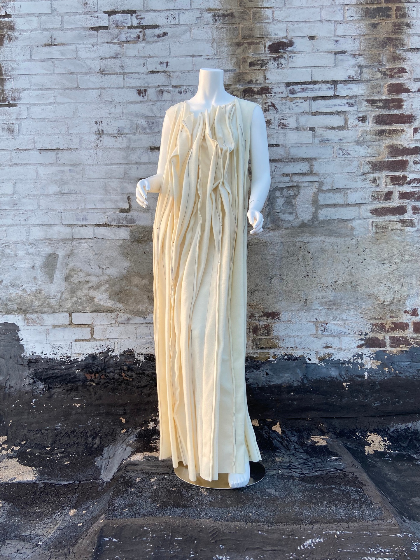 Mannequin wears a floor-length shift dress made of a heavy off-white fabric, constructed with many vertically-running wrinkles and ridges. Mannequin is posed against a dirty whitewashed brick wall.