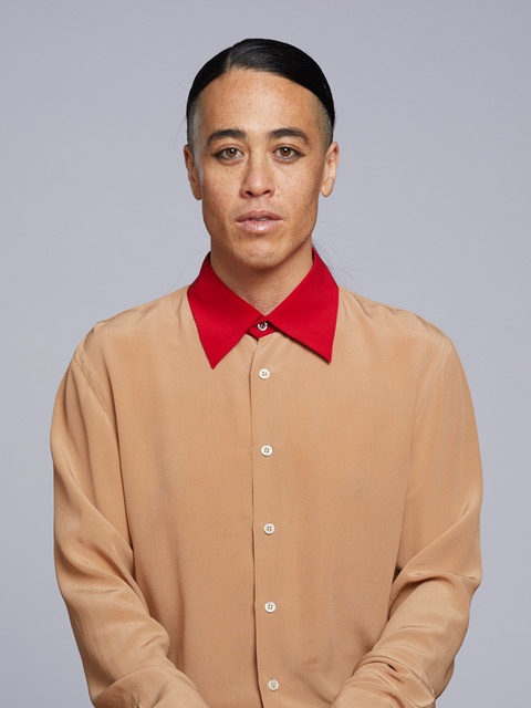 The artist Wu Tsang standing in a tan button-down shirt with a bright red collar buttoned to the top. She wears her dark hair pulled back tight against her head.