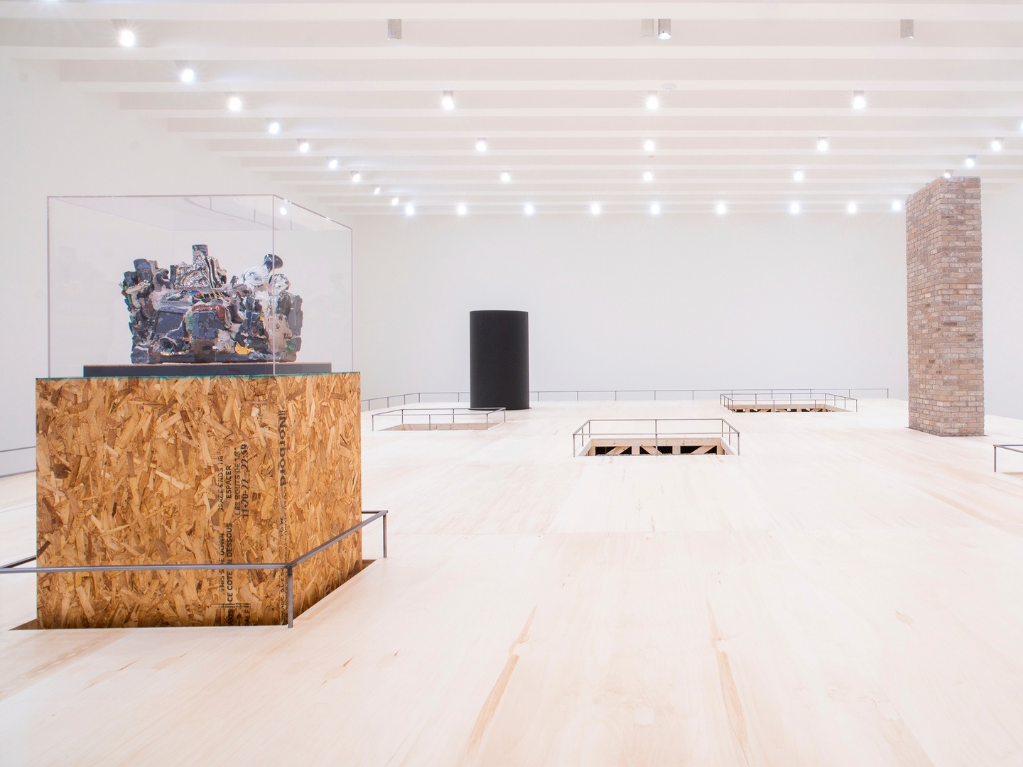 Three large rectangular sculptures, seemingly made of plywood, detritus, bricks, and black stone, set on a wooden platform