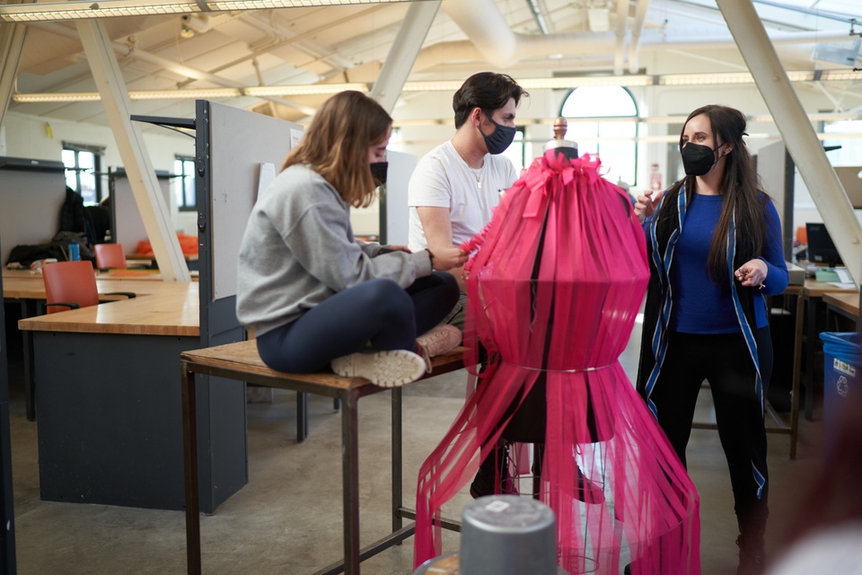 Two students talking with a faculty member, gathered around a large, fuschia fabric garment.