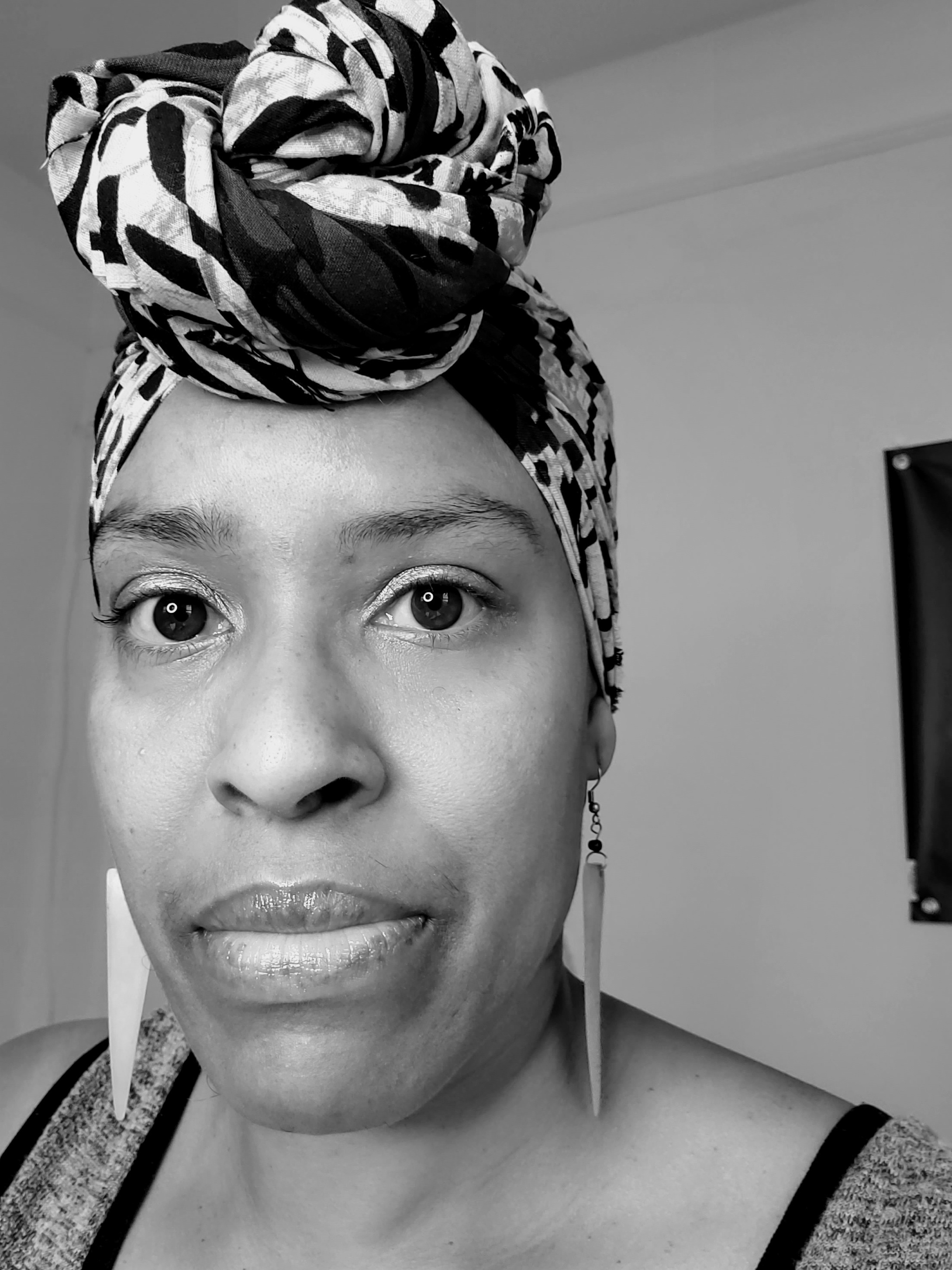 A Black woman looks into the camera with a neutral expression. She wears a headscarf knotted above her forehead and long, narrow triangle-shaped earrings.
