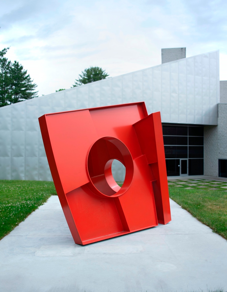 Another view of a large red geometric sculpture sitting in the grass on a concrete platform in front of a gray triangular building.