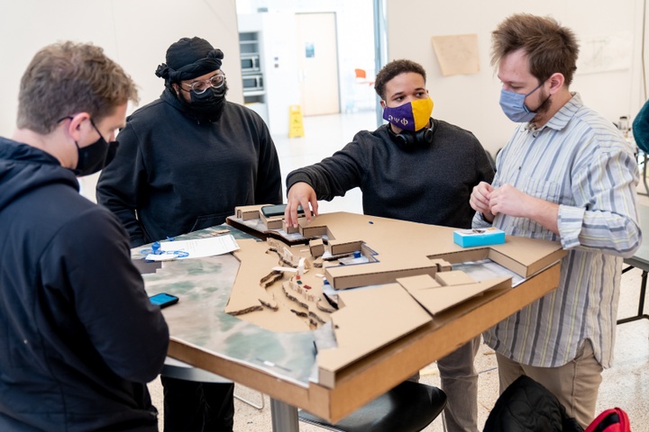 Four students stand around a cardboard city plan model while one gestures at a section of it.