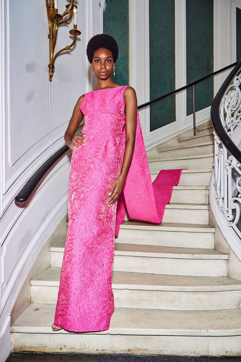 A slender, Black woman stands on a stairway posing in a long, pink gown with a train trailing behind her.