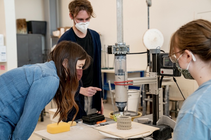 Faculty member Kelley Murphy leans over to watch as a vase is 3D printed, with two students looking on.