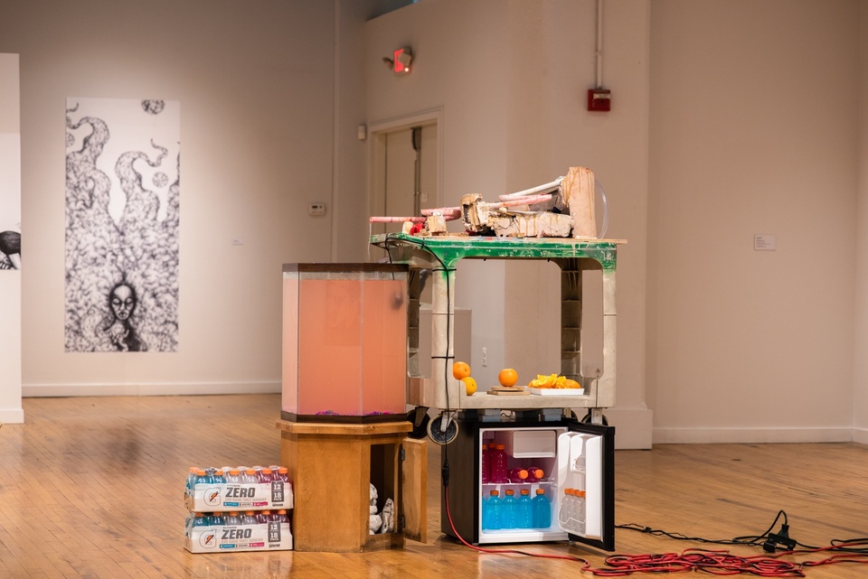 Construction consisting of a minifridge, a plastic wheeled cart, a wooden endtable, a hexagonal aquarium tank, and electrical cables. The fridge is filled with Gatorade bottles, the cart is stacked with oranges, orange slices, and a pile of sticky dirty masonry and tubes. The tankard is full of a murky orange liquid, which is being filled by a trickle from a tube on top of the cart. 