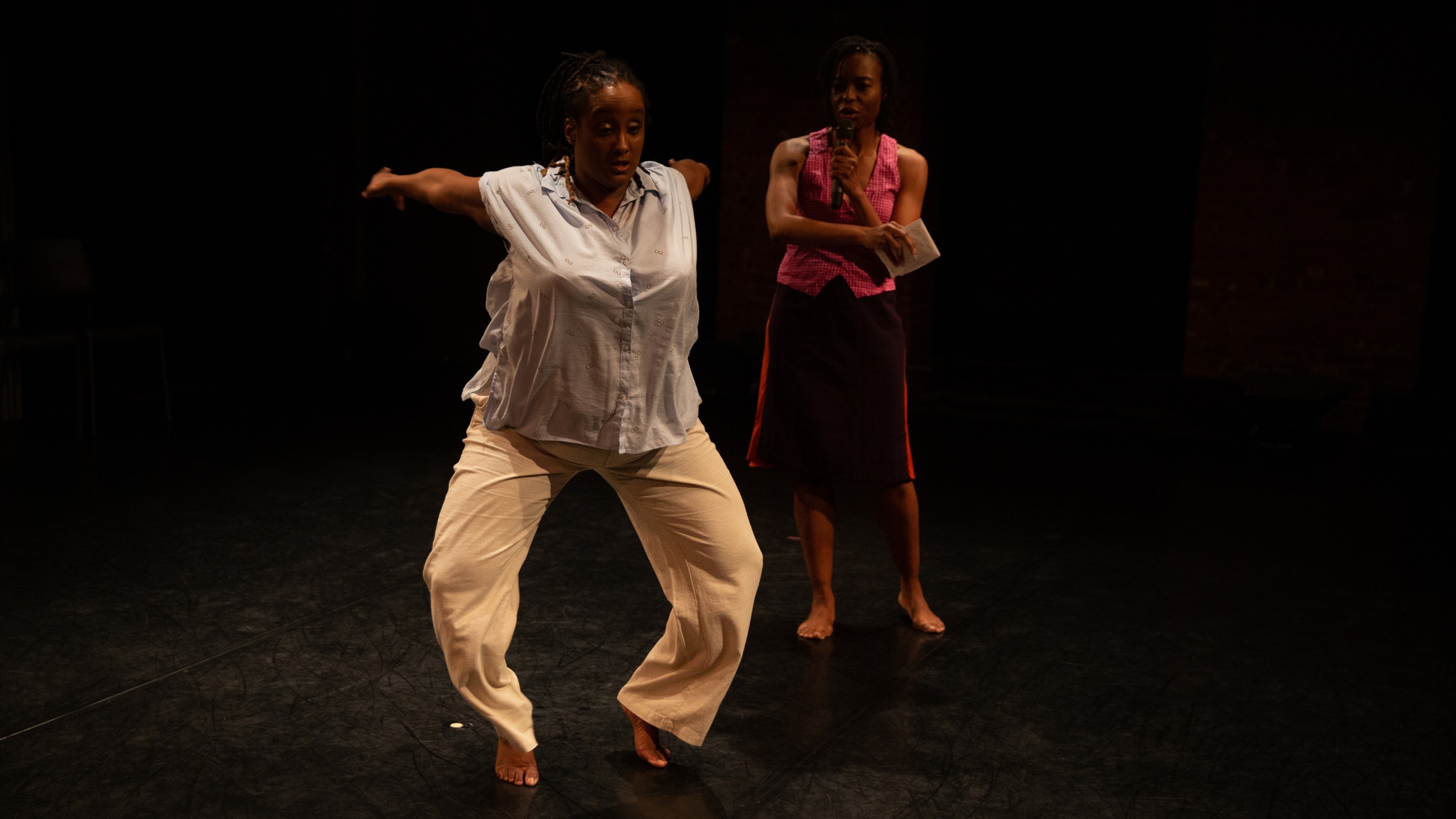 A photo of Kayla Hamilton and Nicole McClam, two dark-skinned Black women. They are both on a stage. Nicole is diagonally behind Kayla. She is wearing dark red mid-length shorts & a pink tank top. She is observing Kayla dancing. Kayla is facing front and has her knees bent as her arms are energetically swung behind her. Kayla is wearing beige pants with a white sleeveless blouse.