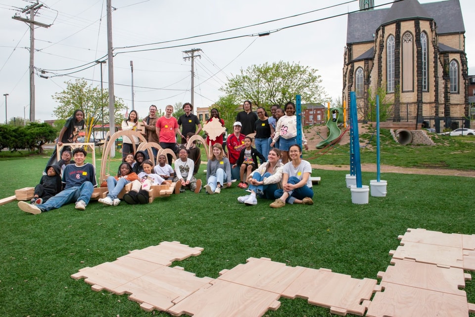 A large group of students and children pose outdoors with modular wooden play structures on a lawn in front of a historic stone church.