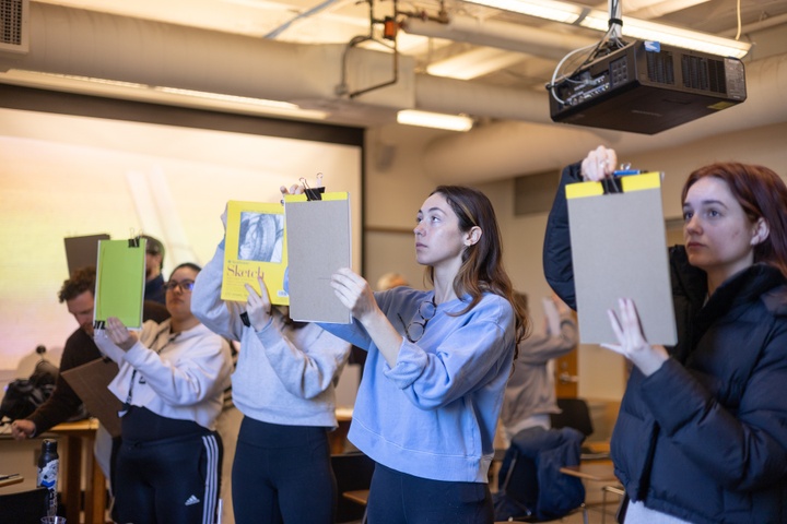 Group of people in a warmly lit room hold up sketchbooks in front of their faces, looking between the page and something projected out of frame in front of them.
