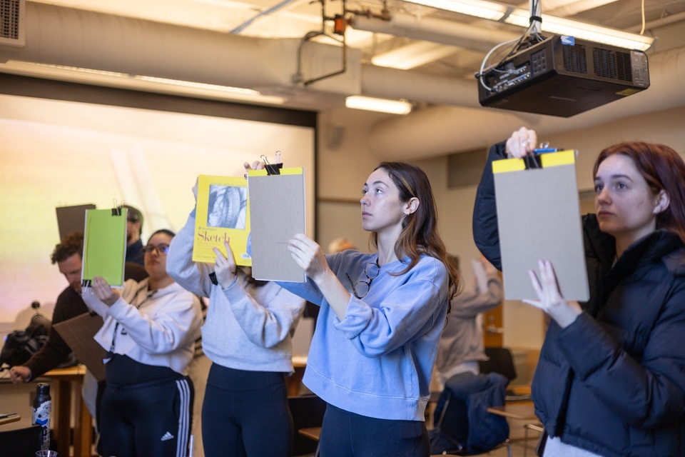 Group of people in a warmly lit room hold up sketchbooks in front of their faces, looking between the page and something projected out of frame in front of them.