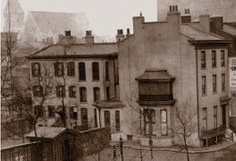 Sepia-toned image of a three-story, 19th-century townhouse on a city corner, with a two-story bay window. In the distance, a church is visible through the fog.