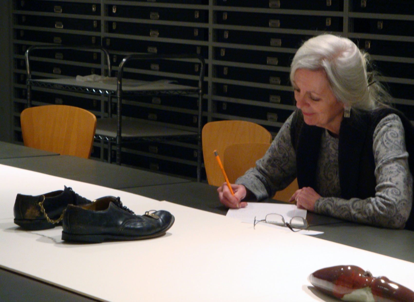 A gray haired woman sits at a table with an artwork on it. She writes in a notebook.