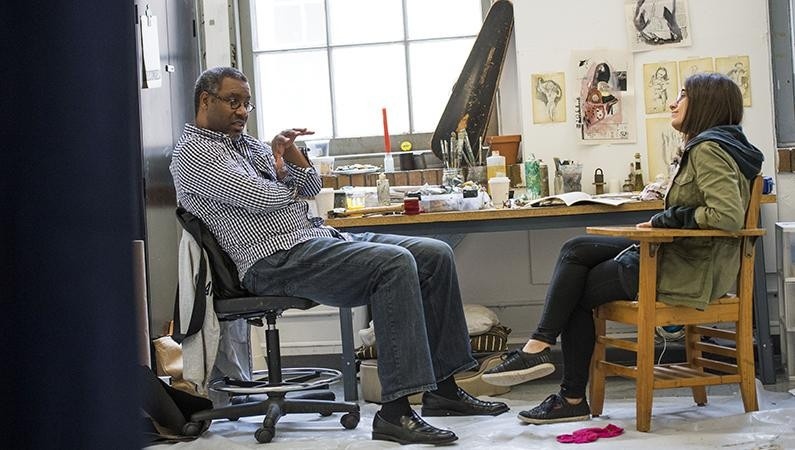 A man in a gingham shirt, blue jeans, and black loafers sits in a chair (at left) talking to a woman (at right) in a green, army-style coat, black hoodie, black jeans, and black sneakers. Daylight illuminates the studio space from a large window at left. Artmaking materials are scattered across a worktable behind the figures. Sketches are pinned to the wall adjacent to the window.