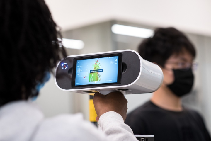 Person points a handheld 3D scanner at another's head, creating a heatmap image of the head on the device's screen.
