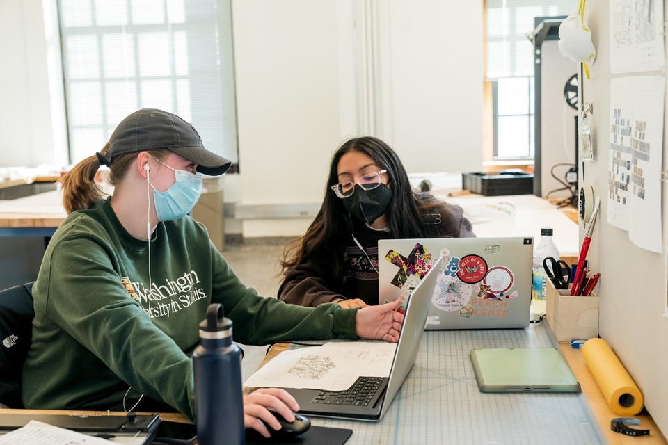 Two smiling people sit together at a desk with their laptops, comparing their screens.