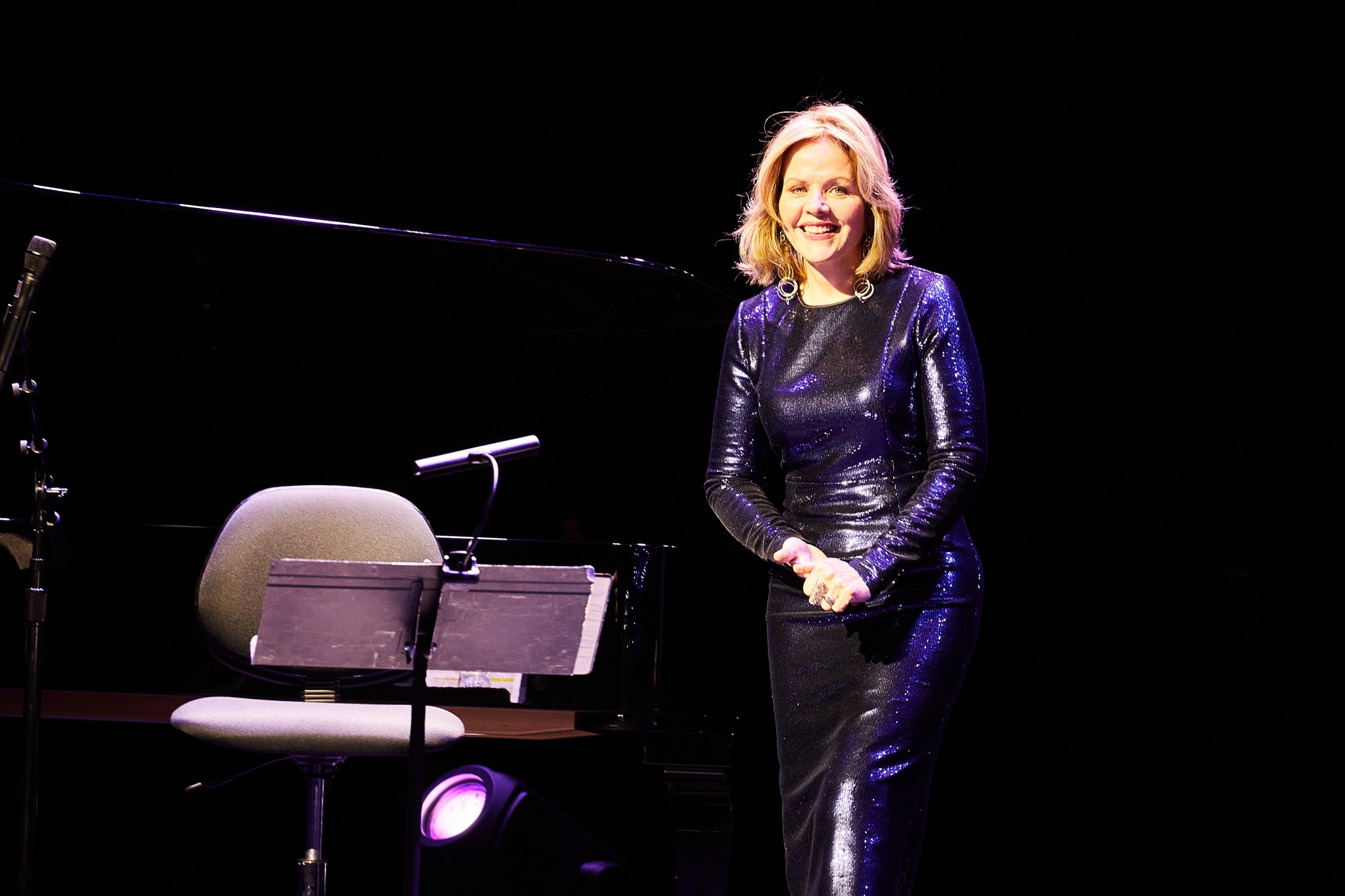 A singer smiling alone on a stage in front of a dark background. She is a white woman who wears a glittery, sequined purple dress and holds her hands together at her waist.