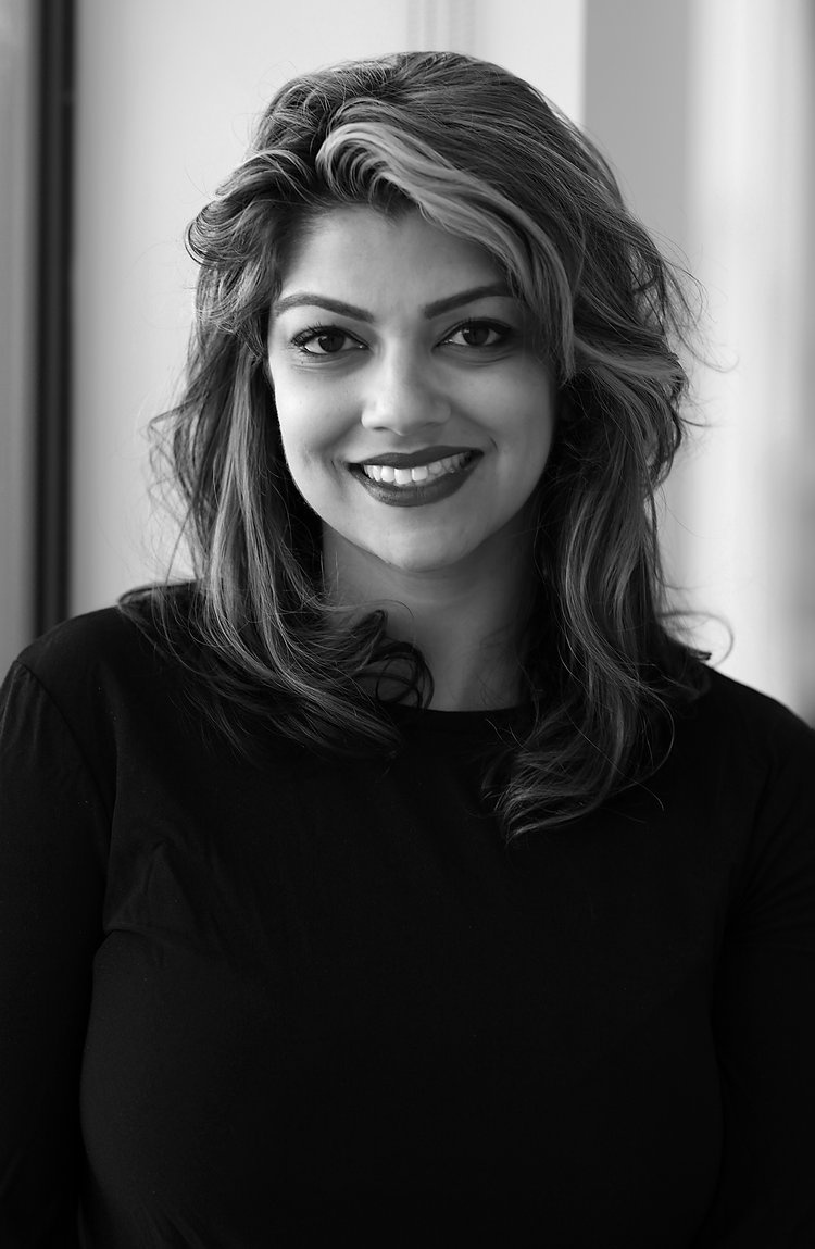 A black-and-white photo of Jasmine Wahi smiling at the camera, her hair falling to her shoulders. 
