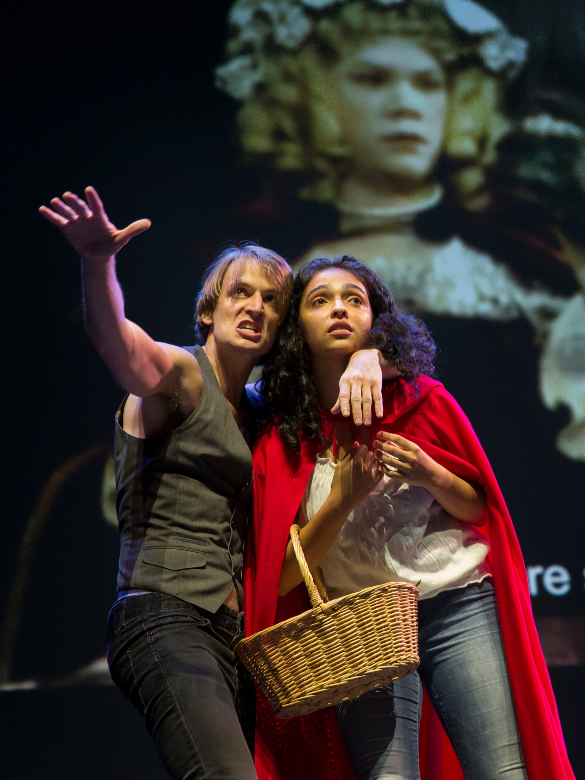 Two Deaf performers, a young man and woman, stand closely together on a stage with a video projection behind them. The white man, wearing a dark gray vest, has one arm around the shoulders and neck of the other performer while stretching his other arm out toward us. The young Hispanic woman, with curly dark hair to her shoulders, wears a red riding hood cloak and holds a basket in the crook of her arm. The video projection shows a period scene from a performance of Into the Woods, with the subtitled words “singing sweetly” visible behind the performers. 