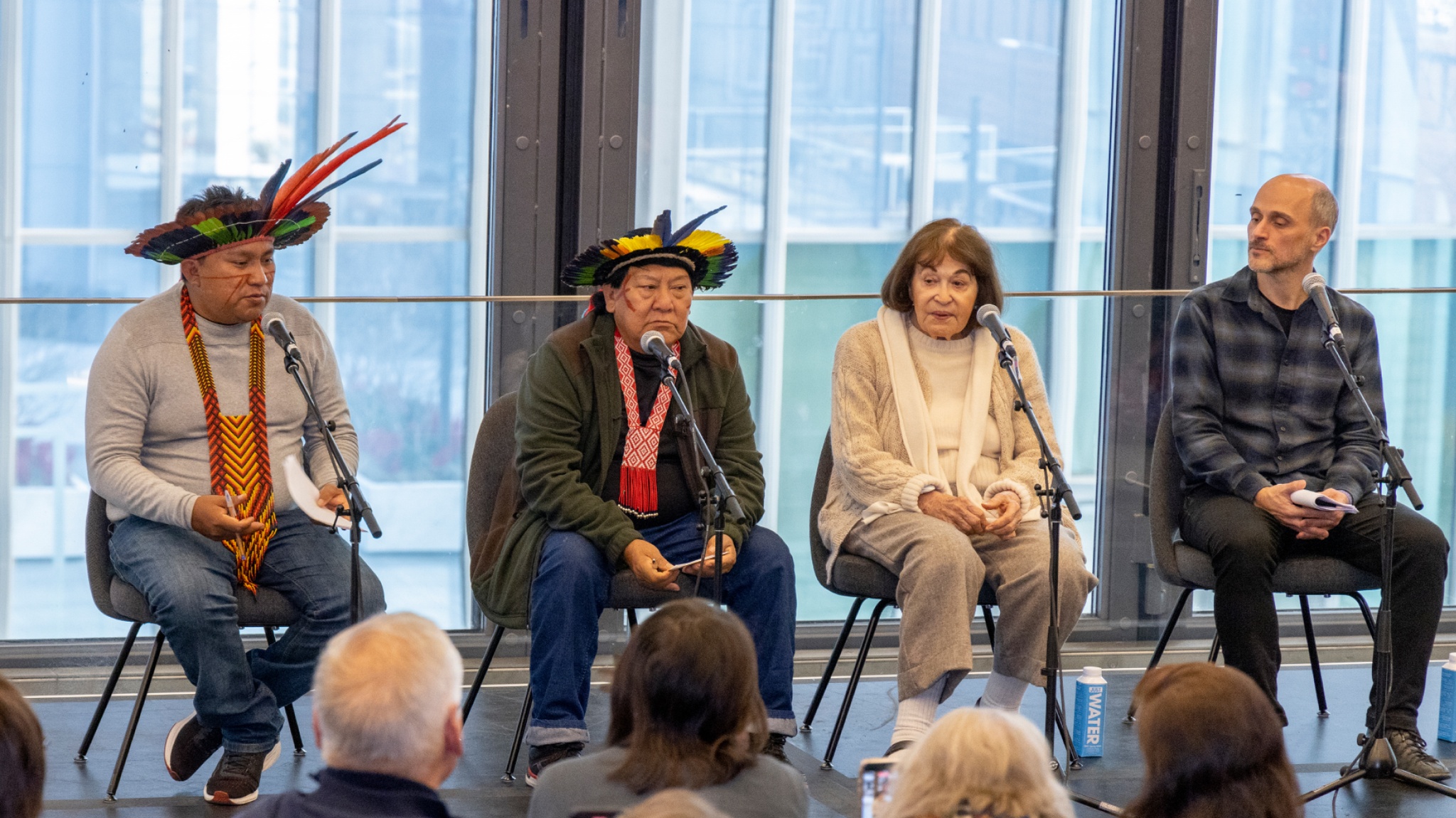 Four panelists sit on a stage with large windows behind them. On the left are Dario and Davi Kopenawa, two Yanomami men who wear head dresses made of colorful feathers. On the right of them sits artists Claudia Andujar, a Brazilian white woman with brown hair wearing an off white outfit. To her right is curator Thyago Nogueira, a white Brazilian man wearing a dark button down shirt in a check plaid pattern.