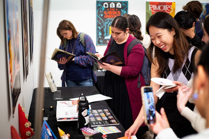 People read copies of illustrated books off a table in a small gallery space displaying posters on the walls above each stack of books.