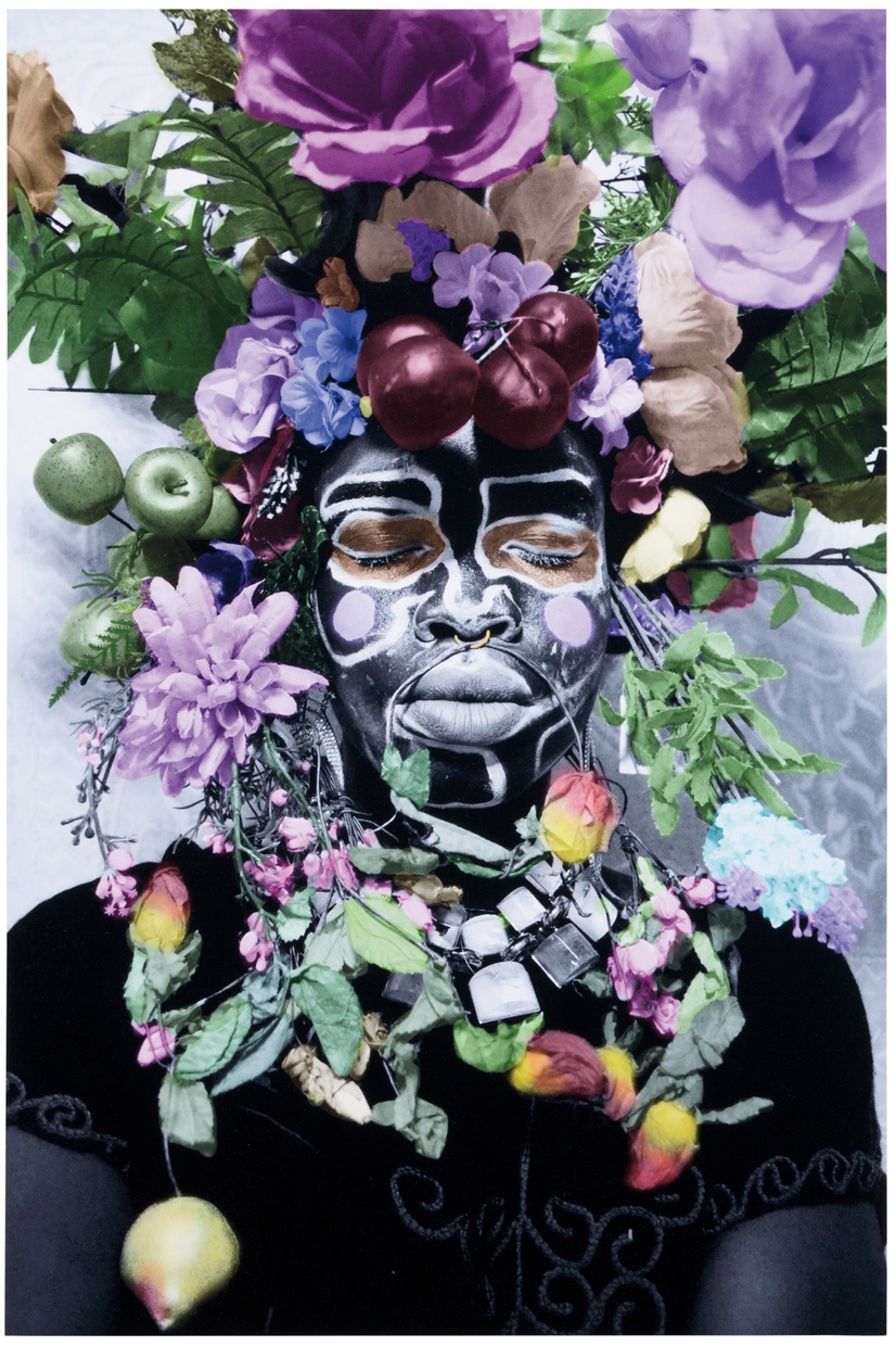 A portrait of a black woman with closed eyes that are ringed by gold and with thick white paint outlining her features. Her face is surrounded by fruits and flowers of various purple, pink, yellow and green hues.