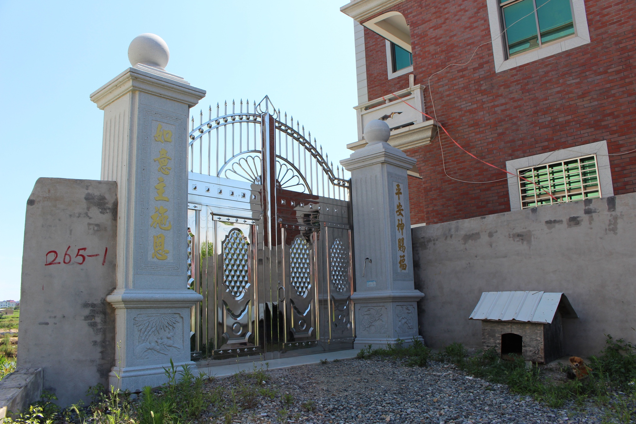Two concrete columns with writing in gold Chinese characters that stand with a steel gate closed between them. To the right rise a concrete wall and brick, two-story house. To the side of the left column, an empty lot with green grass is visible in the background.