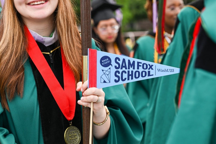 Close up of a smiling graduate in a crowd, wearing a green robe with a medal around their neck, holding a standard and a blue pennant reading "Sam Fox School, WashU23."