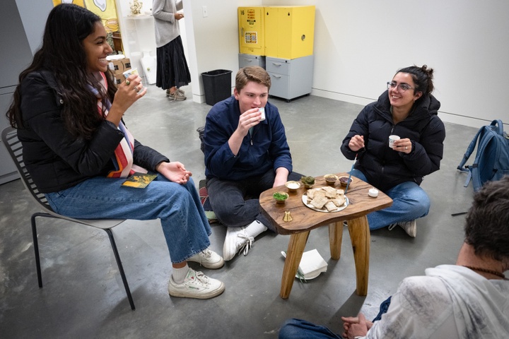 Three people sit around a tiny wooden table and react to something they've just eaten.