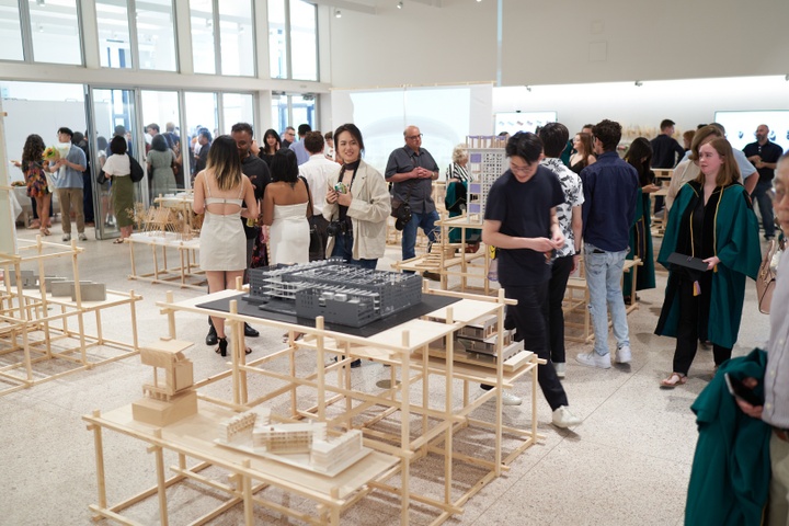 Gallery space arranged with architectural models on tables and projections onto a bank of screens. People mill around the tables looking at the work.