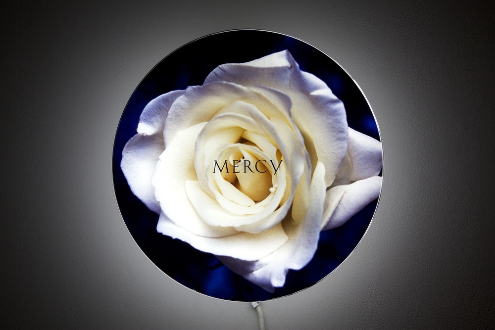 A wall-mounted circular lightbox with the image of a white rose on a dark background and the word "MERCY" in the center.