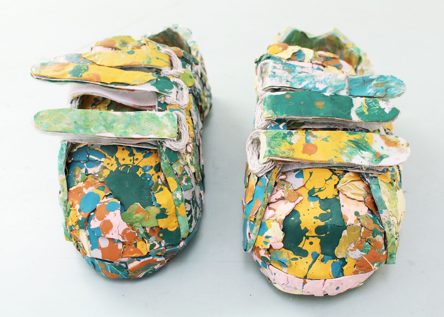 Pair of velcro closure shoes covered in layers of paint blotches in yellow, teal, and shades of pink and clay.